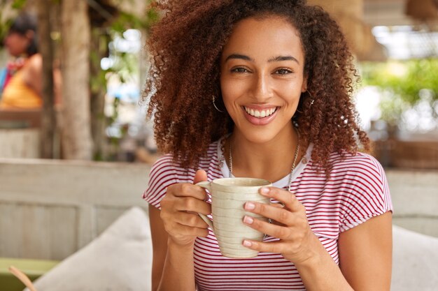 Feliz mujer negra sostiene una taza con bebida aromática caliente, sonríe positivamente, vestida con una camiseta a rayas, tiene el cabello oscuro y nítido, disfruta de una conversación agradable con un amigo en el restaurante