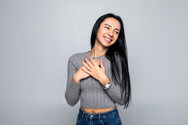 Feliz mujer pacífica aislada en la pared gris del estudio, tome las manos en el pecho cerca del corazón, sonriendo, agradecida, femenina, se siente agradecida, muestra aprecio y amor