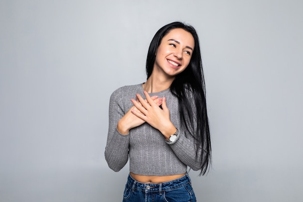 Feliz mujer pacífica aislada en la pared gris del estudio, tome las manos en el pecho cerca del corazón, sonriendo, agradecida, femenina, se siente agradecida, muestra aprecio y amor