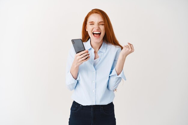 Feliz mujer pelirroja grita de alegría y satisfacción, gana un premio en línea, sostiene un teléfono inteligente y aprieta una mano en señal de triunfo, pared blanca