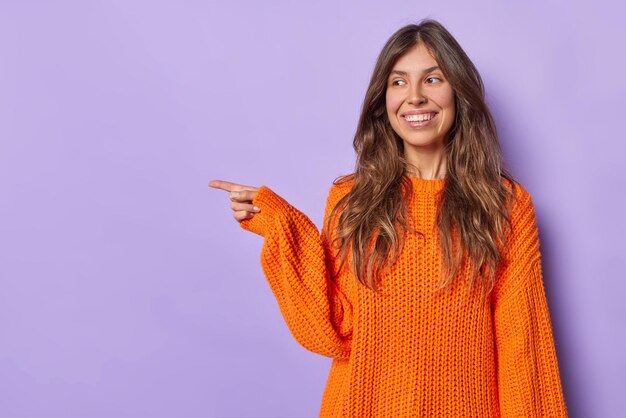 Feliz mujer de pelo largo apunta hacia afuera muestra la dirección de algo vestido con un suéter naranja tejido demuestra el lugar para su anuncio aislado sobre fondo púrpura. Forma de banner de venta