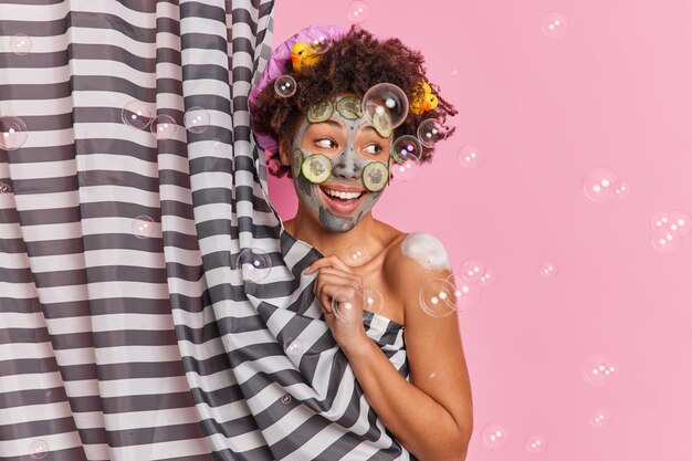 Feliz mujer de pelo rizado aplica máscara de arcilla con rodajas de pepino disfruta de la ducha mira a un lado posa positivamente medio desnuda detrás de la cortina tiene patos de goma en el pelo aislado sobre fondo rosa