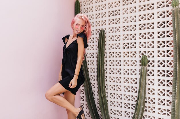 Feliz mujer de pelo rosa posando con sonrisa de sorpresa cerca de cactus.