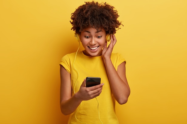 Feliz mujer de piel oscura se siente bien mientras escucha una agradable melodía en auriculares, enfocada en la pantalla del teléfono inteligente