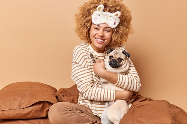 Feliz mujer positiva con el pelo rizado abraza al perro pug expresa amor y cuidado usa pijama de máscara de dormir se queda en la cama junto con un nuevo miembro de la familia aislado sobre fondo marrón Concepto de hora de dormir