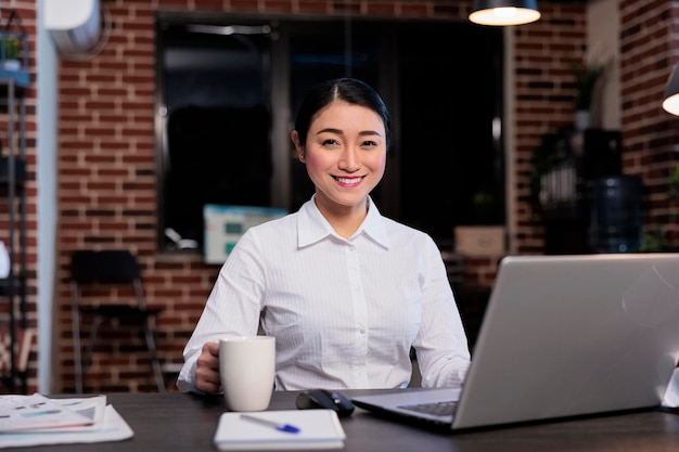 Feliz mujer positiva sentada en el escritorio en el espacio de trabajo de la oficina de la empresa comercial. Gerente ejecutivo atractivo y confiado sentado en el espacio del departamento de marketing mientras sonríe a la cámara.