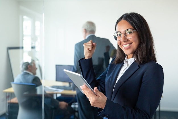 Feliz mujer profesional en gafas y traje sosteniendo tableta y haciendo gesto de ganador mientras dos hombres de negocios trabajan detrás de una pared de vidrio. Copie el espacio. Concepto de comunicación