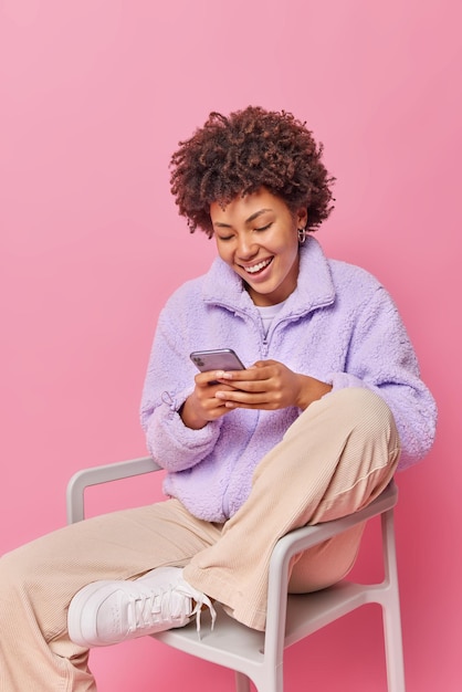 Foto gratuita feliz mujer relajada contenta utiliza información de actualizaciones de teléfonos móviles en el sitio web utiliza conexión gratuita a internet vestida con ropa elegante se sienta en una silla aislada sobre fondo rosa envía mensajes de texto