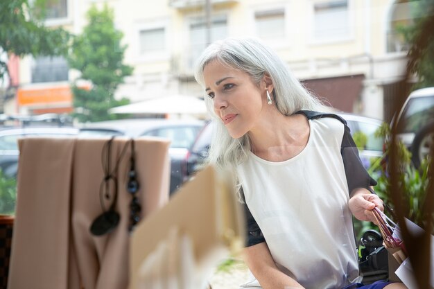 Foto gratuita feliz mujer relajada mirando los accesorios en el escaparate, sosteniendo bolsas de la compra, de pie en la tienda exterior. vista frontal a través del cristal. concepto de escaparate