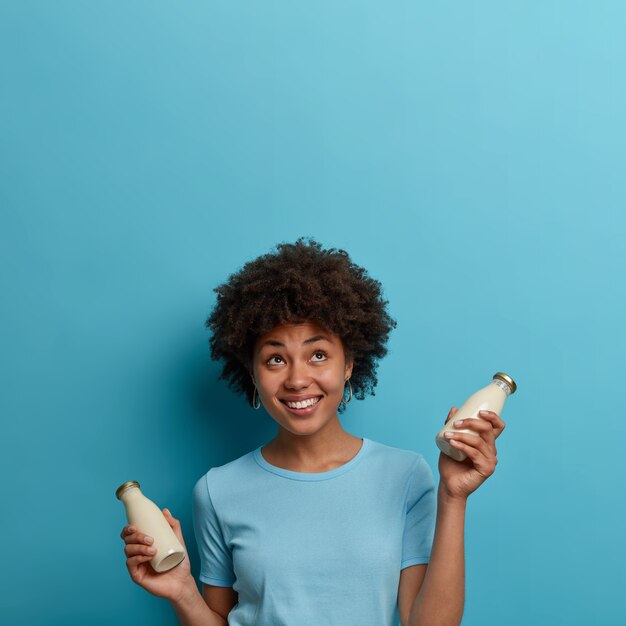 Feliz mujer rizada étnica bebe bebidas sin lactosa, sostiene una botella de leche de almendras o de coco, mira hacia arriba, sonríe positivamente, aislado sobre una pared azul, copie el espacio para su información