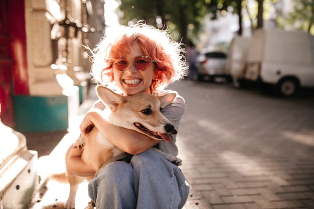 Feliz mujer rizada en jeans sonríe sinceramente y abraza a perro corgi