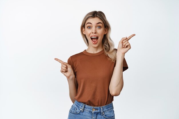 Feliz mujer rubia apuntando con el dedo hacia los lados y sonriendo, mostrando dos opciones de venta, diferentes variantes, de pie en camiseta en blanco