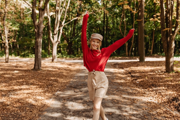 Feliz mujer rubia en un bonito suéter rojo y pantalones cortos beige bailando en el parque de otoño. Mujer joven con estilo posando con alegría en buen tiempo al aire libre.