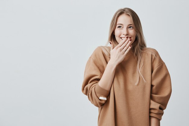 Feliz mujer rubia cerrando la boca con las manos va a ver sorpresa preparada por el marido de pie y sonriendo