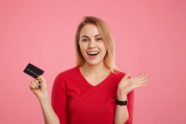 Feliz mujer rubia emocionada tiene tarjeta de plástico, no espera recibir salario, va a hacer pagos, mira alegremente a la cámara