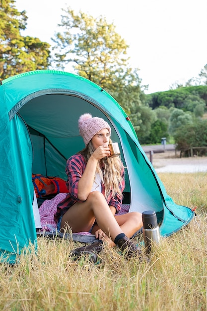 Feliz mujer rubia con sombrero bebiendo té, sentada en la tienda y mirando a otro lado. Viajero caucásico de pelo largo acampar en el césped en el parque y relajarse en la naturaleza. Concepto de turismo, aventura y vacaciones de verano.