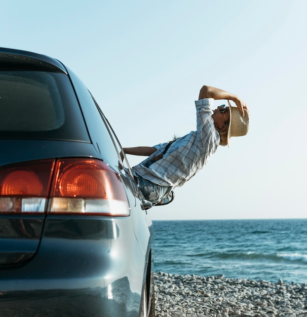 Feliz mujer rubia con sombrero de pie fuera de la ventana del coche