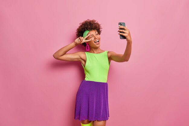 Feliz mujer saliente hace gesto de victoria, guiña el ojo y posa en la cámara del teléfono inteligente, toma selfie, vestida con traje colorido