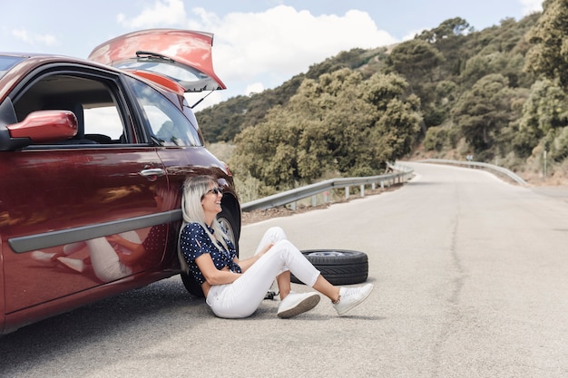 Feliz mujer sentada cerca del coche averiado en la carretera sinuosa