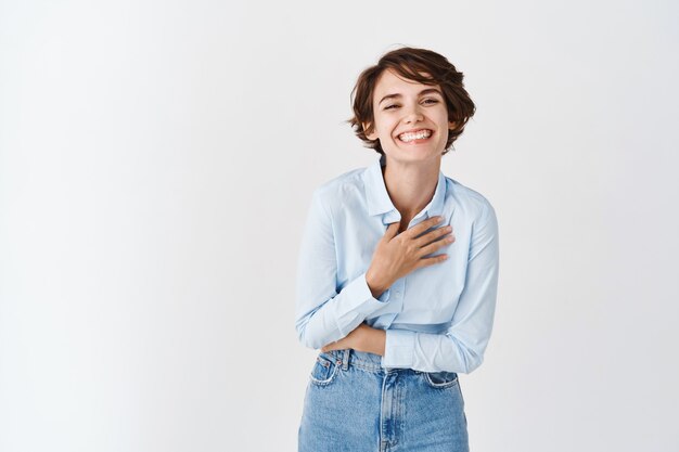 Feliz mujer sincera riendo y sonriendo sin preocupaciones, tocando el pecho mientras se ríe entre dientes sobre una broma divertida, de pie en la pared blanca