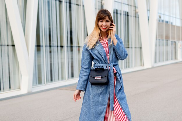 Feliz mujer sonriente en abrigo azul de moda y vestido rojo a rayas caminando en el moderno centro de negocios