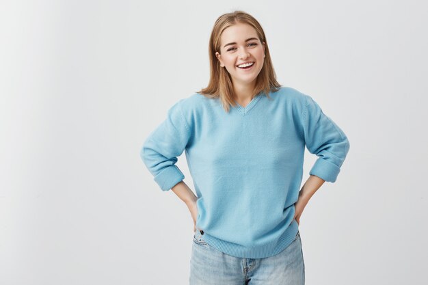 Feliz mujer sonriente con apariencia atractiva y cabello rubio con suéter azul y jeans que muestran su amplia sonrisa, que tiene buen humor y le gusta posar, poniendo las manos en las caderas.