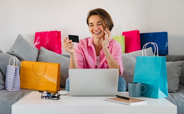 Feliz mujer sonriente en camisa rosa en el sofá en casa entre coloridas bolsas de la compra con tarjeta de crédito pagando en línea en la computadora portátil