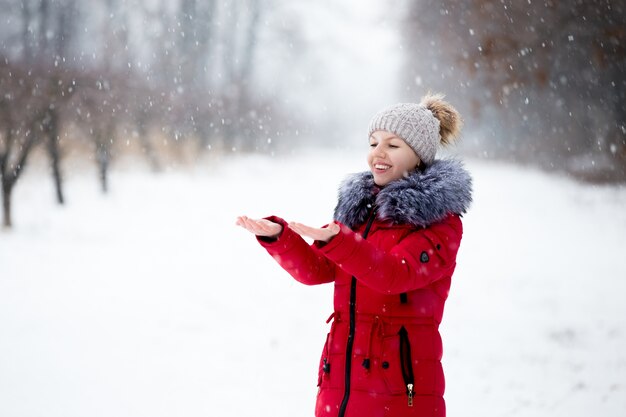 Feliz mujer sonriente en chaqueta de invierno rojo coger la nieve con sus palmas, al aire libre, en el parque