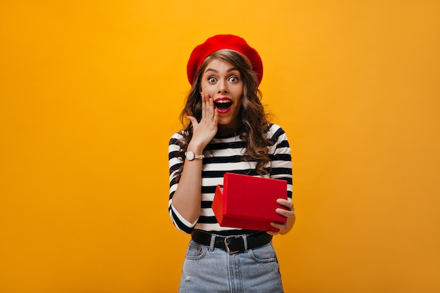 Feliz mujer sorprendida en boina roja tiene caja de regalo. Dama sorprendida con cabello ondulado con sombrero brillante y camisa a rayas se regocija.