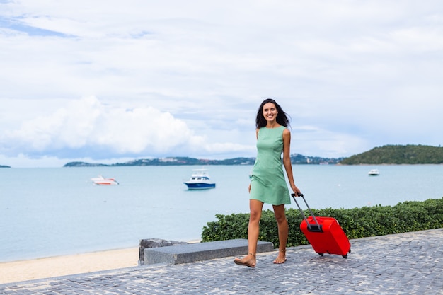 Feliz mujer turista elegante pelo bastante largo caucásico vestido con maleta roja fuera del hotel