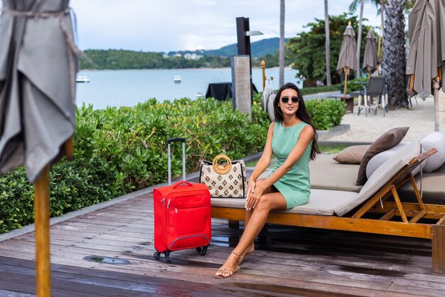 Feliz mujer turista elegante pelo bastante largo caucásico vestido con maleta roja fuera del hotel