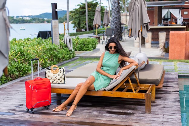 Feliz mujer turista elegante pelo bastante largo caucásico vestido con maleta roja fuera del hotel