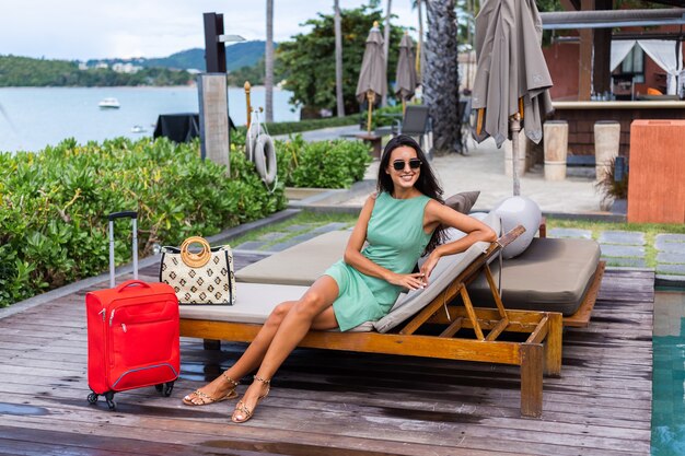 Feliz mujer turista elegante pelo bastante largo caucásico vestido con maleta roja fuera del hotel