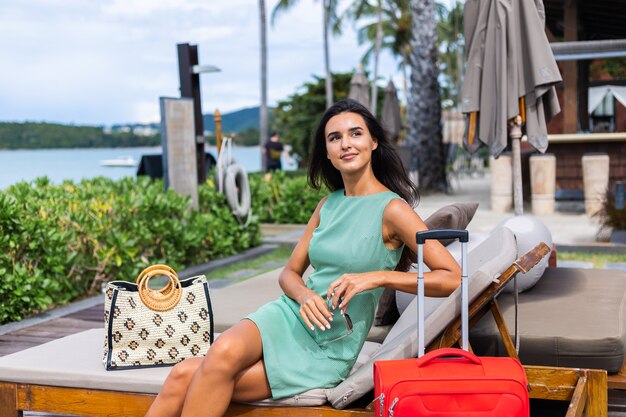 Feliz mujer turista elegante pelo bastante largo caucásico vestido con maleta roja fuera del hotel