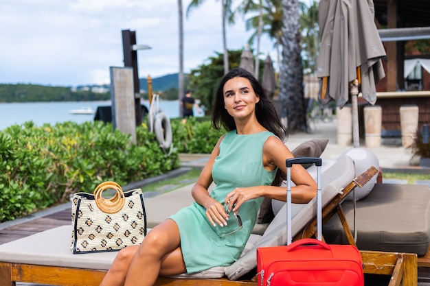 Feliz mujer turista elegante pelo bastante largo caucásico vestido con maleta roja fuera del hotel