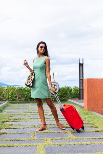 Feliz mujer turista elegante pelo bastante largo caucásico vestido con maleta roja fuera del hotel