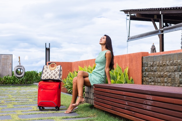 Feliz mujer turista elegante pelo bastante largo caucásico vestido con maleta roja fuera del hotel