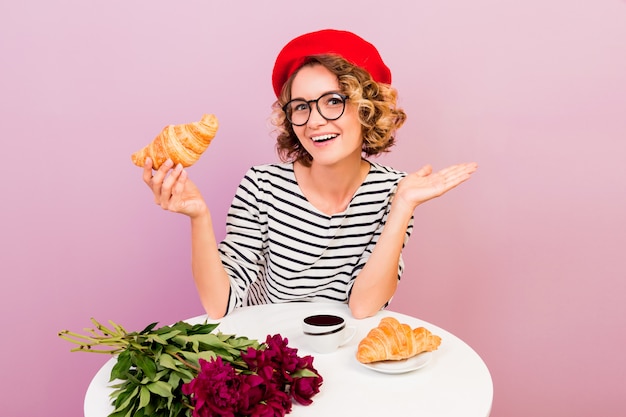 Feliz mujer viajera en Francia comiendo croissans con café, se sienta junto a la mesa en rosa.