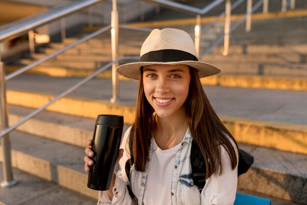 Feliz mujer viajera con mochila y sombrero sosteniendo termo