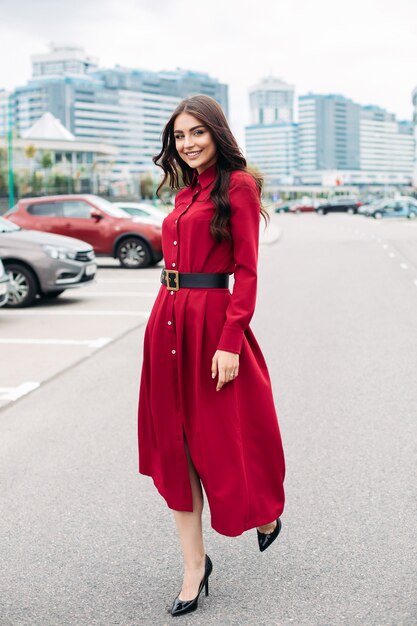 Feliz muy jovencita en vestido rojo mirando a la cámara mientras camina por la calle en la ciudad moderna. Concepto de estilo de vida