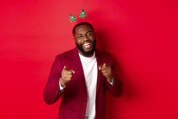 Feliz Navidad. Apuesto hombre afroamericano en blazer y diadema de fiesta, celebrando el año nuevo, apuntando a la cámara y sonriendo, felicitando, fondo rojo