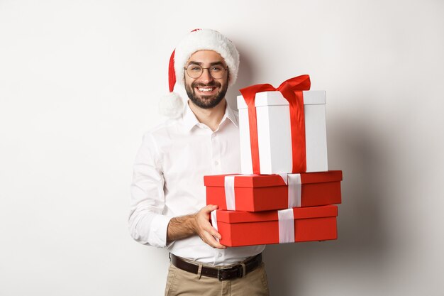 Feliz navidad, concepto de vacaciones. Feliz joven sonriendo, sosteniendo regalos en cajas y vistiendo gorro de Papá Noel