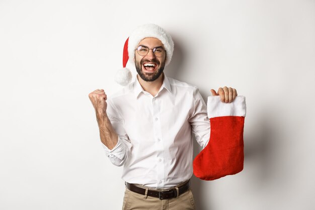 Feliz navidad, concepto de vacaciones. Hombre adulto feliz recibe regalos en calcetín de Navidad, mirando emocionado, vistiendo gorro de Papá Noel