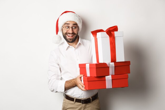 Feliz navidad, concepto de vacaciones. Hombre emocionado celebrando la Navidad, vistiendo gorro de Papá Noel y sosteniendo regalos, de pie