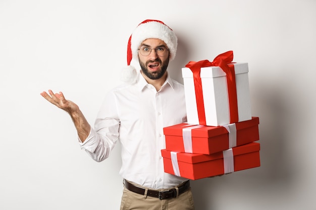 Feliz navidad, concepto de vacaciones. Hombre que parece confundido mientras sostiene los regalos de Navidad, encogiéndose de hombros perplejo, de pie en el sombrero de santa contra el fondo blanco.