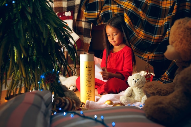 Feliz Navidad y Felices Fiestas. La niña linda del niño escribe la carta a Santa Claus cerca del árbol de Navidad en casa interior. vacaciones, infancia, invierno, concepto de celebración