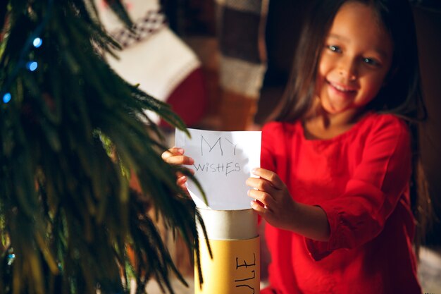 Feliz Navidad y Felices Fiestas. La niña linda del niño escribe la carta a Santa Claus cerca del árbol de Navidad en casa interior. vacaciones, infancia, invierno, concepto de celebración