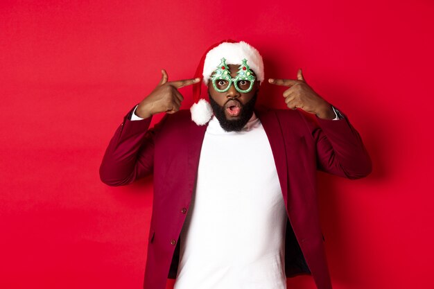 Feliz Navidad. Hombre negro alegre con gafas de fiesta divertidas y gorro de Papá Noel, sonriendo alegre, celebrando las vacaciones de invierno, de pie sobre fondo rojo.