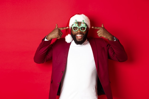 Feliz Navidad. Hombre negro alegre con gafas de fiesta divertidas y gorro de Papá Noel, sonriendo alegre, celebrando las vacaciones de invierno, de pie sobre fondo rojo.