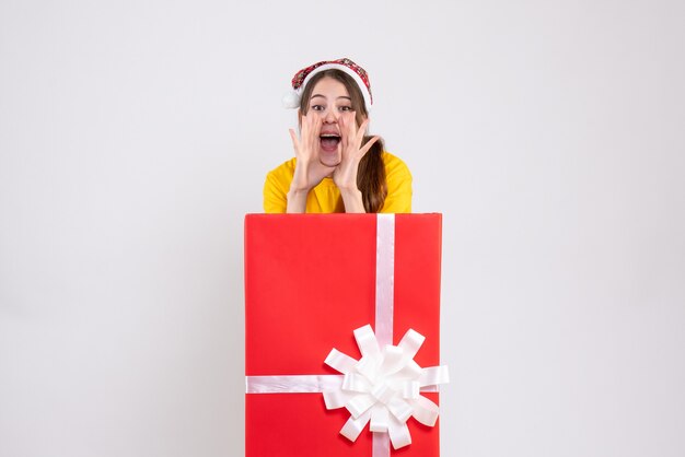 Feliz Navidad niña con gorro de Papá Noel gritando de pie detrás de gran regalo de Navidad en blanco